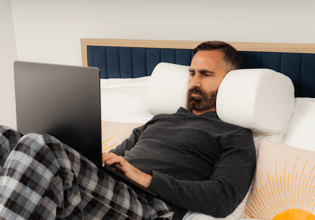 Man relaxing upright in bed with a laptop on the Restinator pillow